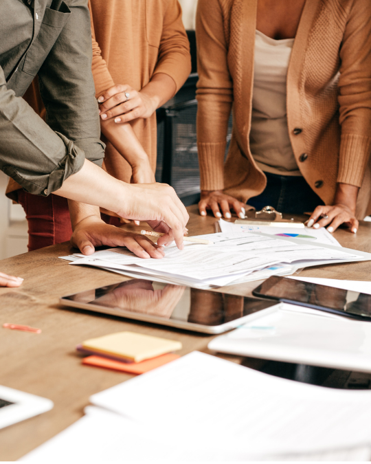 Image of a group of people standing around a table collaboratively assessing printed documents and an iPad